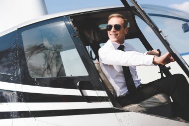 good-looking Pilot in formal wear and sunglasses sitting in helicopter cabin clipart