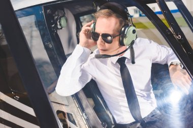 Pilot in sunglasses and headset with microphone sitting in helicopter cabin clipart