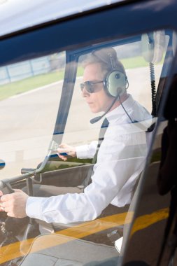 mature Pilot in sunglasses and headphones with microphone sitting in helicopter cabin clipart