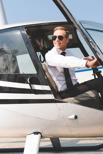 smiling Pilot in sunglasses and formal wear sitting in helicopter cabin and holding wheel