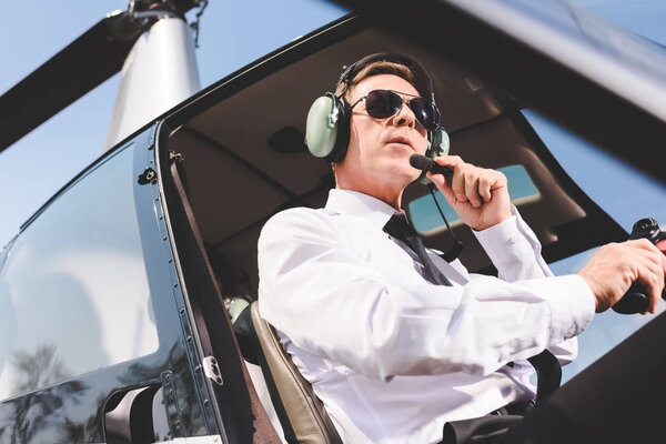 Pilot in sunglasses, formal wear and headset sitting in helicopter cabin