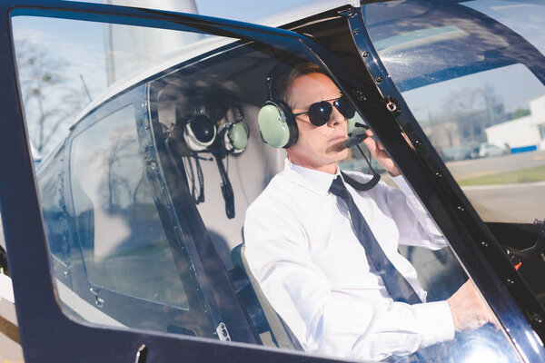 handsome mature Pilot in sunglasses and headset sitting in helicopter cabin