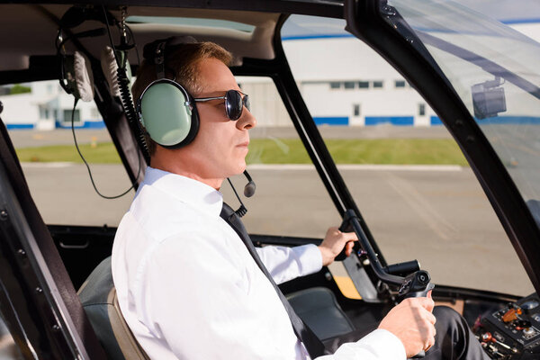 mature Pilot in sunglasses and headset sitting in helicopter cabin and holding wheel