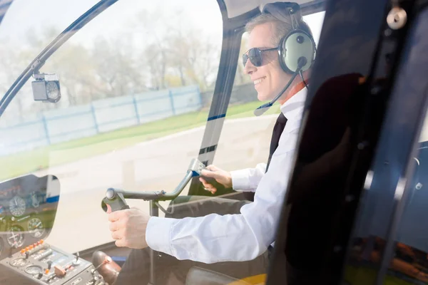 坐在直升机舱里时戴着太阳镜和耳机的成熟飞行员带着麦克风微笑 — 图库照片