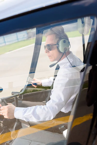 戴着太阳镜和耳机的成熟飞行员 带麦克风坐在直升机机舱里 — 图库照片