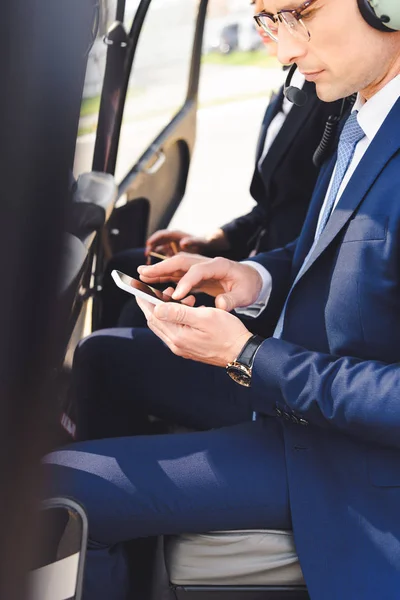 businessman in formal wear sitting in helicopter cabin and using smartphone