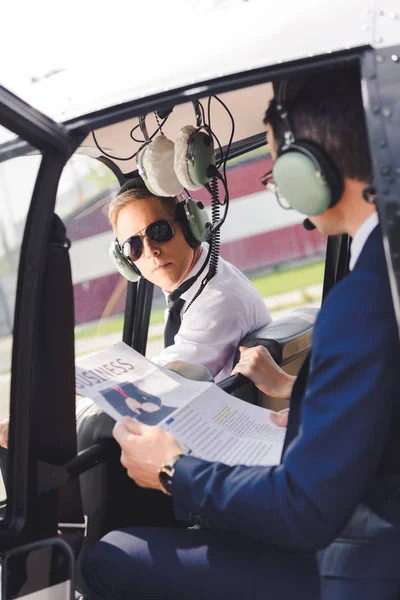 飞行员在耳机和商人与报纸在直升机舱 — 图库照片