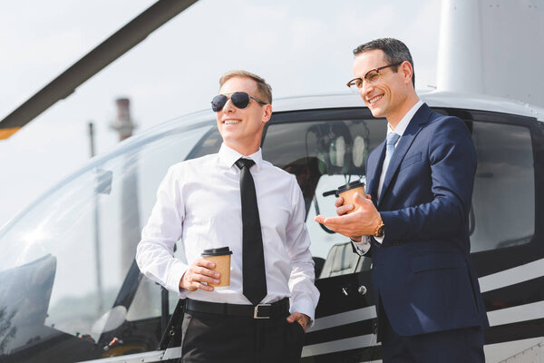 smiling Pilot in sunglasses and businessman with coffee to go near helicopter