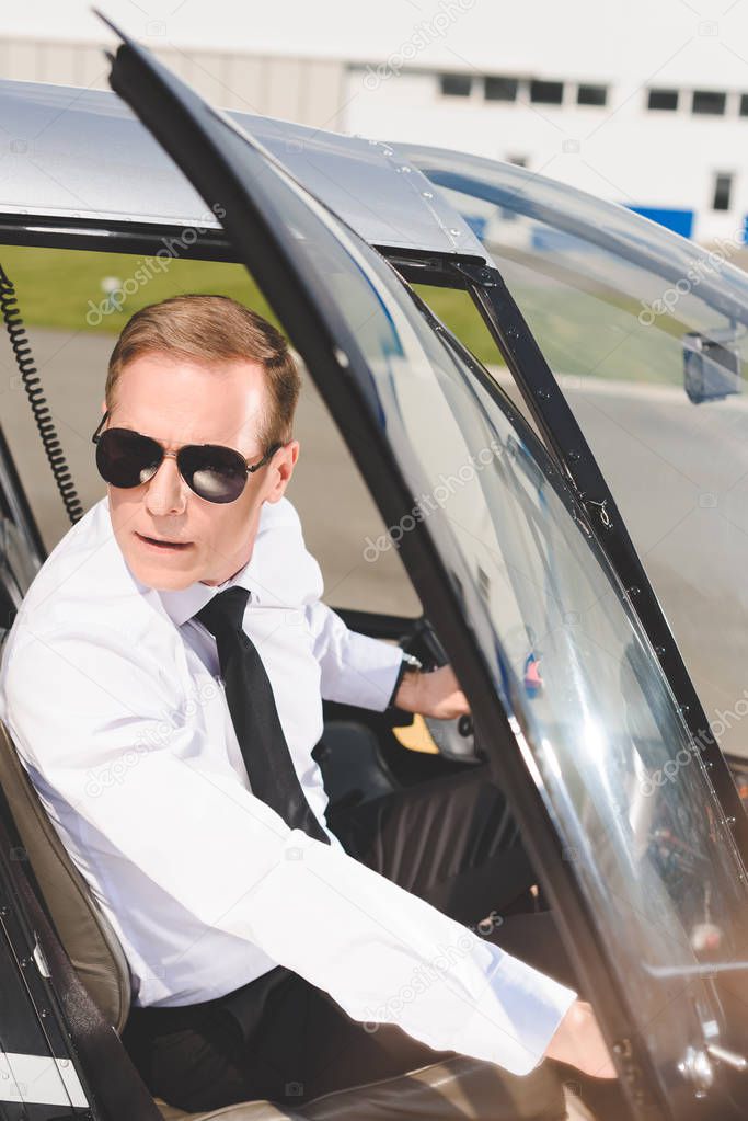 handsome Pilot in sunglasses and formal wear sitting in helicopter cabin and opening door