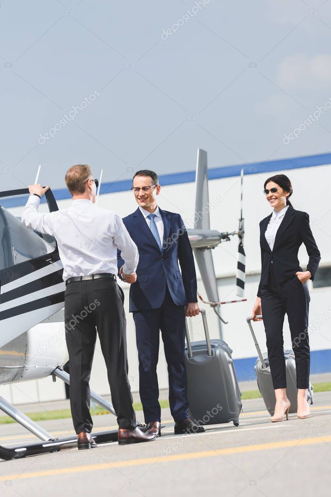 businessman with luggage shaking hands with pilot near helicopter and businesswoman