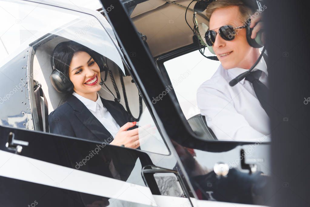 smiling businesswoman and pilot in headsets sitting in helicopter cabin