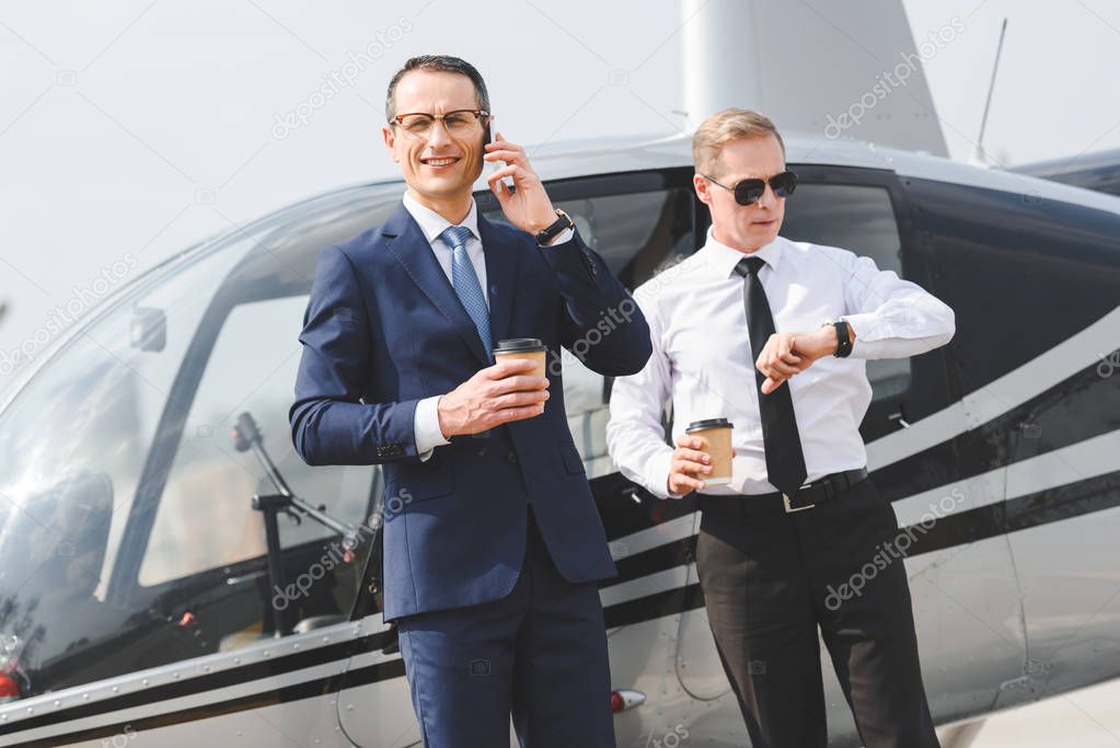 businessman in suit with coffee to go talking on smartphone while pilot looking at watch near helicopter