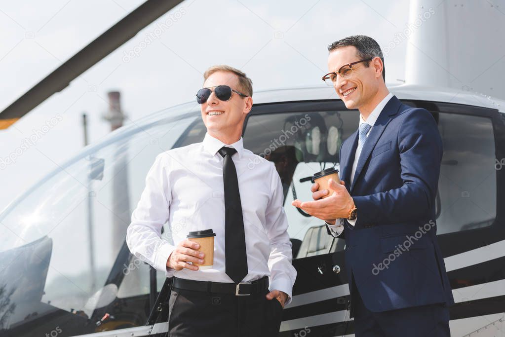 smiling Pilot in sunglasses and businessman with coffee to go near helicopter