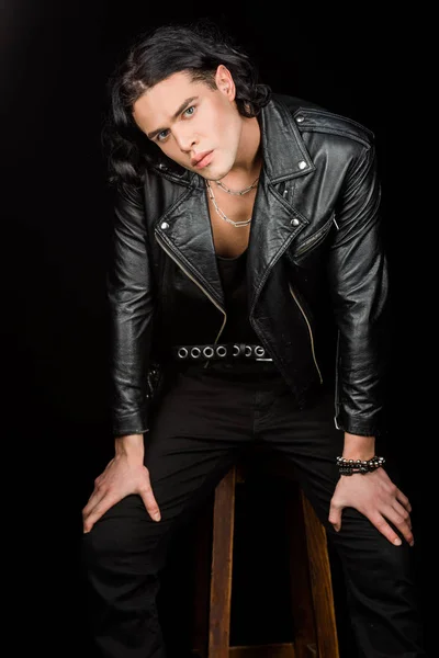 handsome man in leather jacket looking at camera while sitting on chair isolated on black