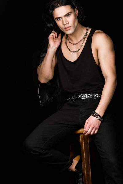 красивый мужчина держит кожаную куртку и смотрит в камеру, сидя на стуле изолированы на черном
 