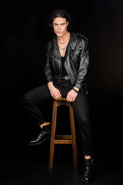 serious man in leather jacket looking at camera while sitting on chair isolated on black
