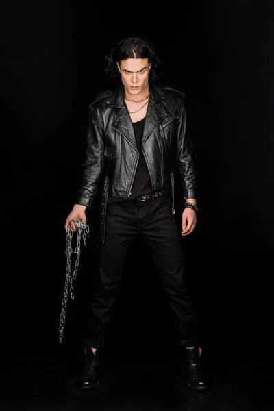 confident man looking at camera and holding chains while standing isolated on black