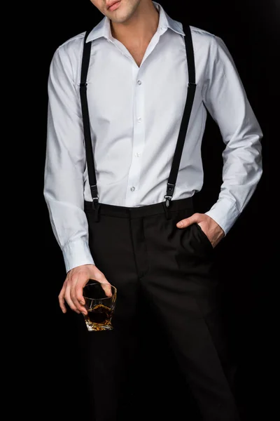 白いシャツを着た男と 黒で隔離されたポケットに手を入れて立ちながらウイスキーのグラスを持つサスペンダーのトリミングされた眺め — ストック写真