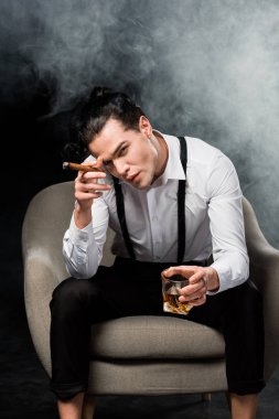 yakışıklı adam kameraya bakıyor ve duman ile siyah viski ve puro cam tutan 