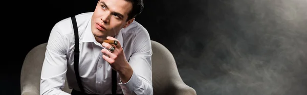 煙で黒いアームチェアに座っている間 葉巻を持っている自信のある男のパノラマショット — ストック写真