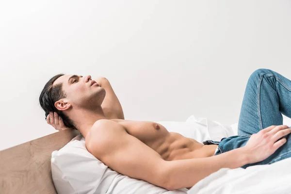 自宅でベッドに横たわっている青いジーンズのハンサムなシャツレスの男 — ストック写真