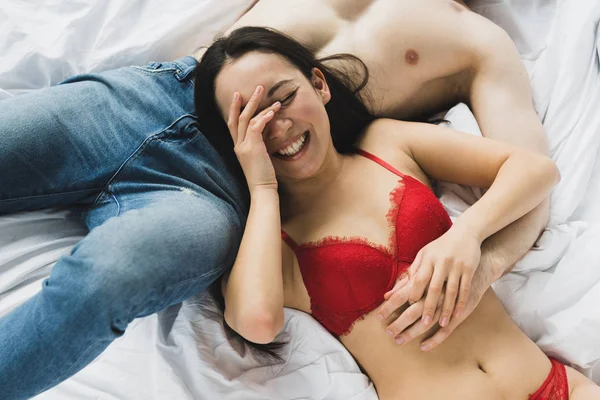 笑うアジアのガールフレンドの近くにベッドに横たわっている青いジーンズのシャツのない男のトリミングされたビュー — ストック写真
