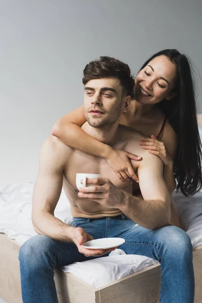Smiling Asian Woman Hugging Pensive Boyfriend Sitting Bed Cup Coffee — Stock Photo, Image