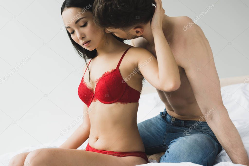 shirtless young man kissing neck of beautiful girlfriend in red lingerie