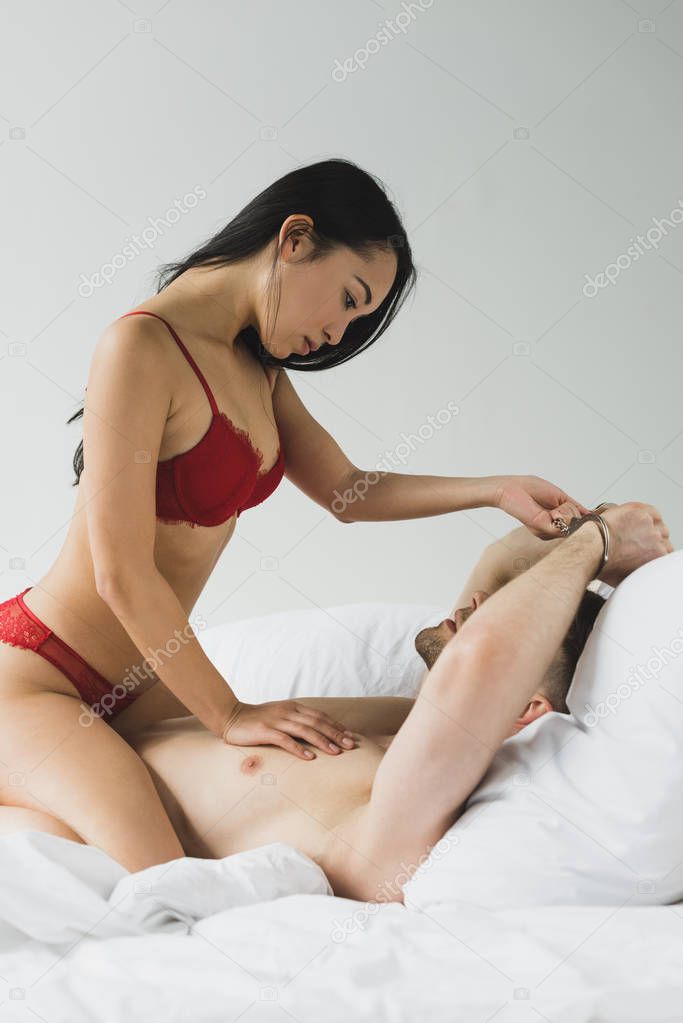 passionate asian woman in red lingerie and shirtless man in handcuffs on white bedding