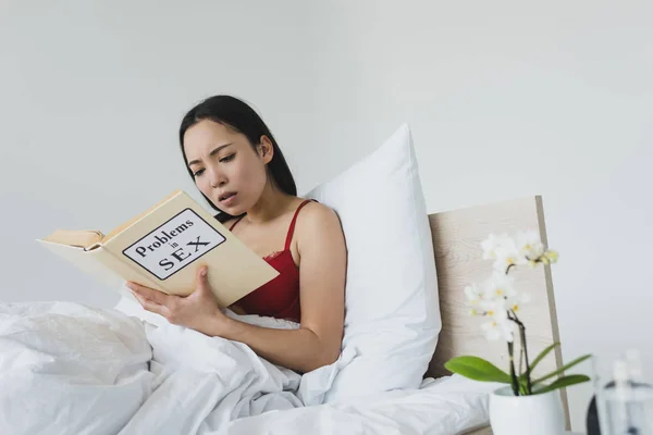 surprised asian woman reading problems in sex book while lying in bed