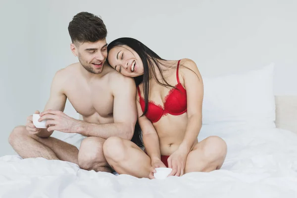 Щаслива Міжрасова Пара Сидить Ліжку Схрещеними Ногами Тримає Чашки Кави — стокове фото