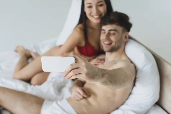 ベッドに横たわって スマートフォンで自分撮りを取る下着で陽気な異人種間のカップル — ストック写真