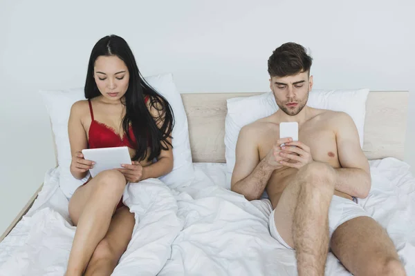 Young Interracial Couple Using Digital Tablet Smartphone While Lying Bed — 스톡 사진