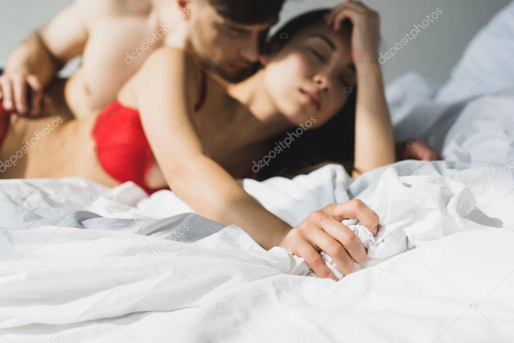 selective focus of man kissing sexy asian girlfriend on white bedding in sunshine