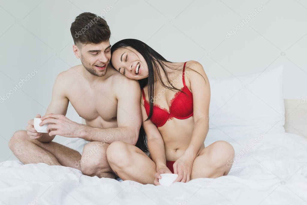 happy interracial couple sitting on bed with crossed legs and holding coffee cups 