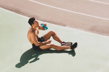 mixed race sportsman sitting at stadium and drinking water clipart