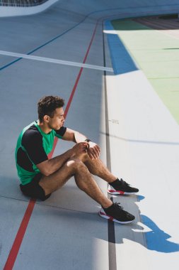 stadyumda koşu pistinde oturan ve SmartWatch 'a bakarak yakışıklı karışık yarış sporcusu 