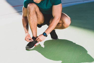 cropped view of sportsman lacing up sneakers, standing on green floor clipart