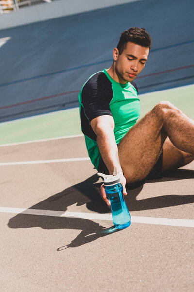 mixed race sportsman sitting at stadium and holding sport bottle with water