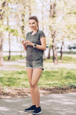 akıllı telefon tutarken ve kulaklıkla müzik dinlerken kameraya gülümseyen çekici sporcu