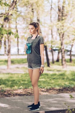 spor şişe tutan spor giyim güzel kadın ve parkta uzağa bakıyor