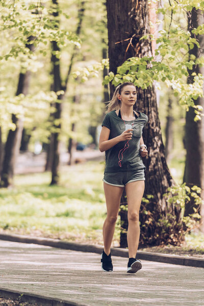 pretty young sportsman running and park, holding smartphone and listening music in earphones