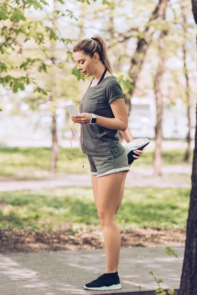 Szép Sportoló Nyújtás Parkban Miközben Használ Smartphone Hallgató Zene Fülhallgató — Stock Fotó