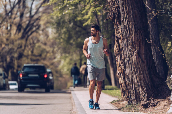 KYIV, UKRAINE - APRIL 25, 2019: Handsome sportsman running along pavement near roadway and listening music in earphones. 