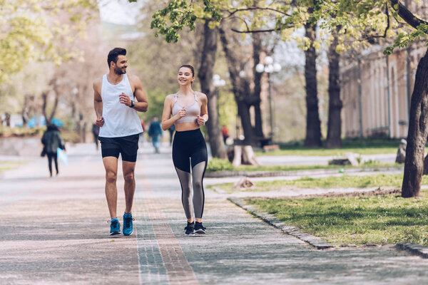 молодой спортсмен и спортсменка улыбаясь во время пробежки вдоль аллеи парка
