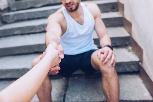 cropped shot of woman giving hand to injured sportsman sitting on stairs and suffering from pain