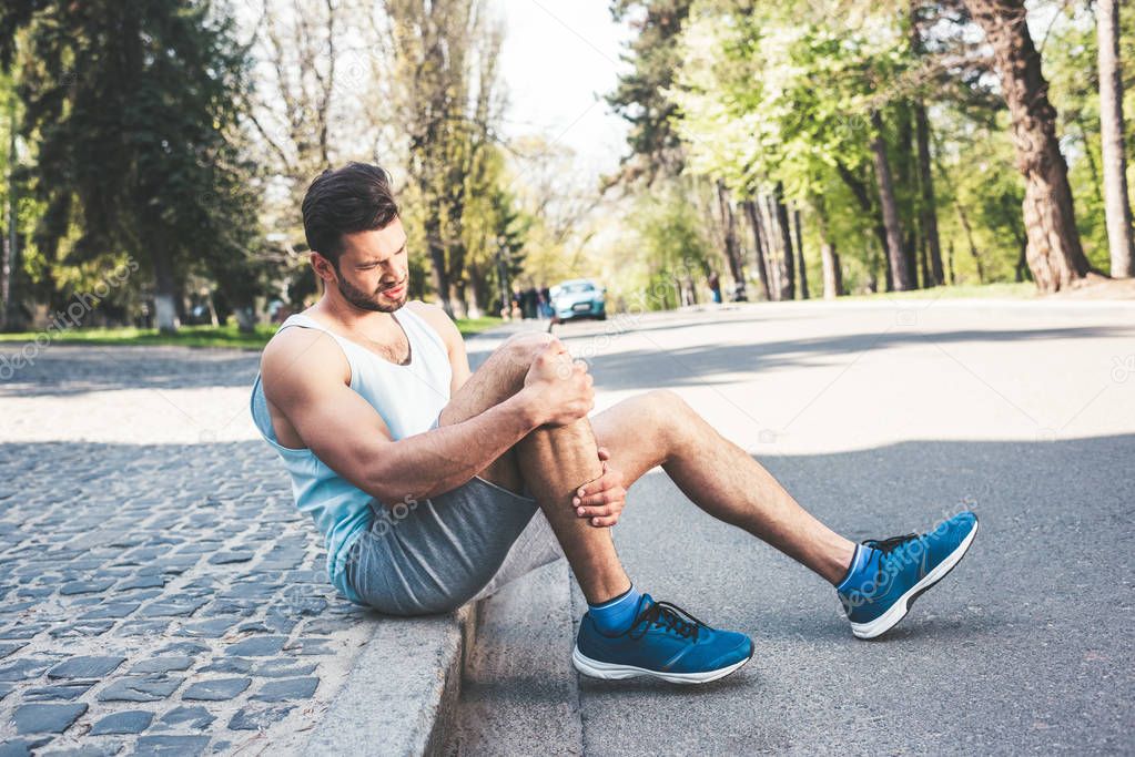 young sportsman sitting on pavement border and touching injured leg