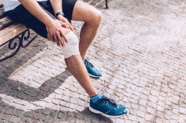 parkta bankta oturan ve yaralı dizine elastik bandaj dokunan sporcunun kısmi görünümü