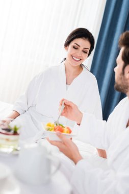 lezzetli meyve salatası yakınında çatal tutan sakallı adam bakarak mutlu kadının seçici odak 