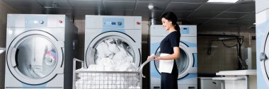 panoramic shot of happy maid looking at dirty bedding while standing near metallic cart  clipart